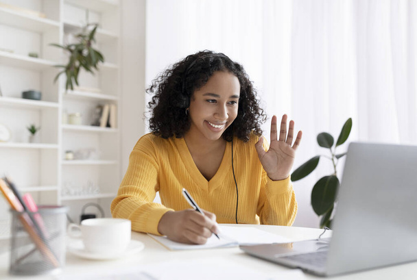 Mulher negra sentada à mesa, usando laptop, tendo conferência virtual, acenando na webcam, cumprimentando o professor em casa - Foto, Imagem