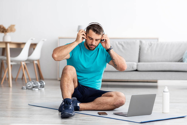 Serious millennial atleta masculino europeu coloca fones de ouvido senta-se no tapete na sala de estar interior com garrafa - Foto, Imagem