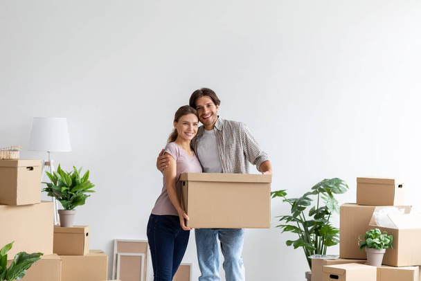 Smiling happy millennial caucasian man and woman with cardboard box hugging in their own apartment - Foto, Imagen