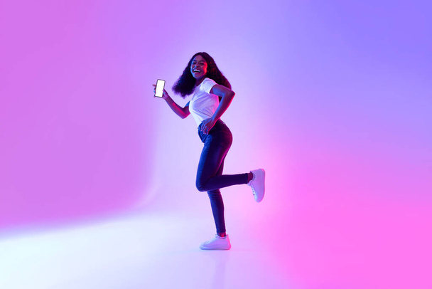 Donna afroamericana allegra con smartphone in esecuzione in luce al neon, mostrando mockup per app mobile o design del sito Web - Foto, immagini