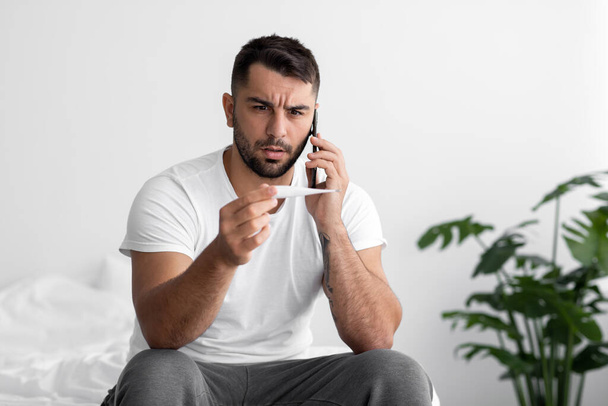 Unhappy european millennial guy suffers from health problems, looks at thermometer, call by phone sit on bed - Foto, imagen
