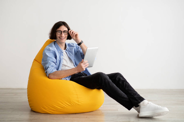 Szczęśliwy młody człowiek za pomocą tabletu cyfrowego podczas siedzenia w fotelu worek na lekkiej ścianie z wolnej przestrzeni - Zdjęcie, obraz