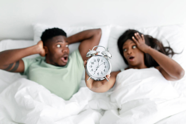 Shocked millennial black male and female with open mouths show alarm clock, lies on white bed in bedroom - Foto, imagen