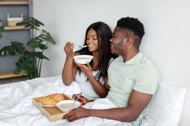 Happy millennial black wife and husband eat porridge on white bed in bedroom, enjoy breakfast and good morning - Photo, Image