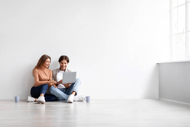 Feliz jovem caucasiano masculino e feminino sentado no chão, descanso, planejando novo interior no computador no quarto vazio - Foto, Imagem