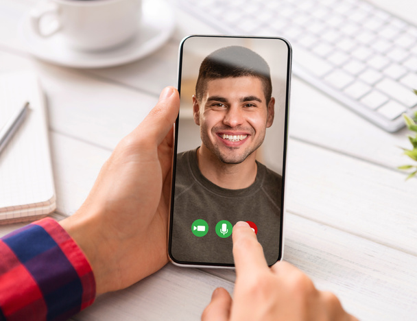 Чоловік має відеодзвінок з другом, сидить на робочому місці і тримає мобільний телефон з хлопцем фото на екрані, колаж
 - Фото, зображення
