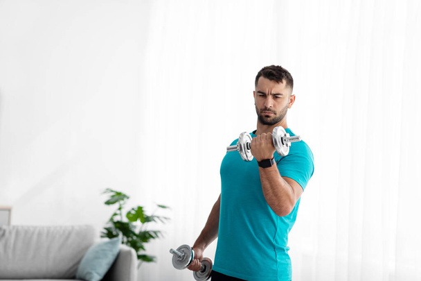 Serious young european guy doing strength exercises for arms, lifting dumbbells in room interior, on window background - Foto, afbeelding