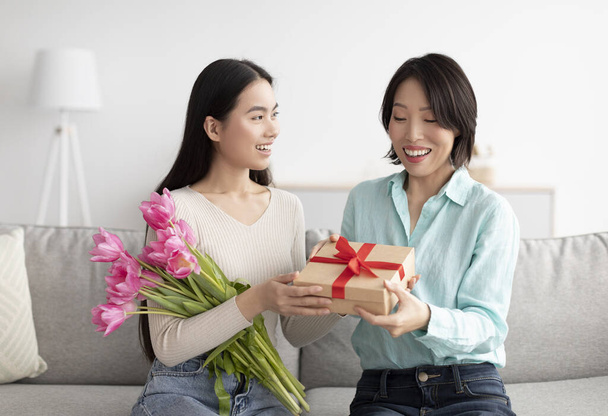 Happy adult Asian daughter giving her mature mom bouquet of flowers and gift box, greeting her with Womans Day at home - Zdjęcie, obraz