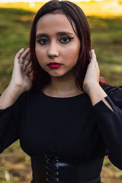 Joven chica de pelo oscuro caucásico con rayas rojas en traje de vestido negro con cinturón de corsé posando para sesión de belleza femenina - Foto, imagen