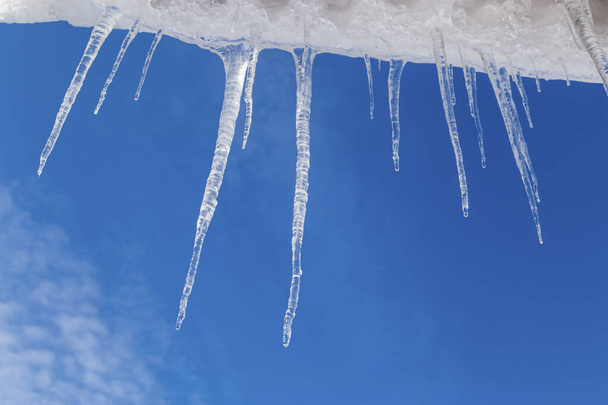 Läpinäkyviä jääpuikkoja sinistä taivasta vasten. Kirkas jää paistaa auringossa talvipäivänä. Jäätynyttä vettä. Jääpuikot, vesipisarat. - Valokuva, kuva