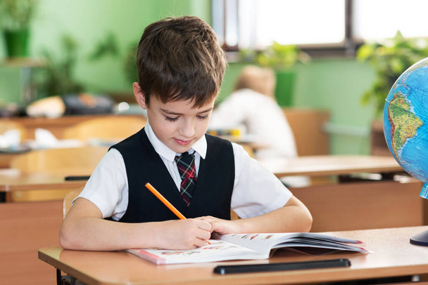 A schoolboy is sitting at a desk in an empty classroom and is writing something in a notebook. School life - Photo, image