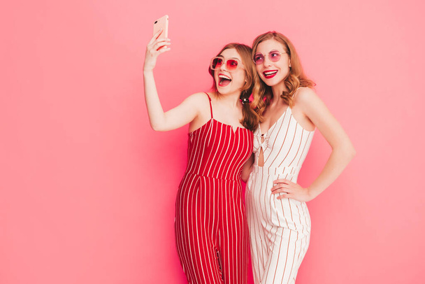 Two young beautiful smiling hipster female in trendy summer overalls clothes. Sexy carefree women posing near pink wall in studio. Trendy and positive models having fun. Taking selfie photos - 写真・画像