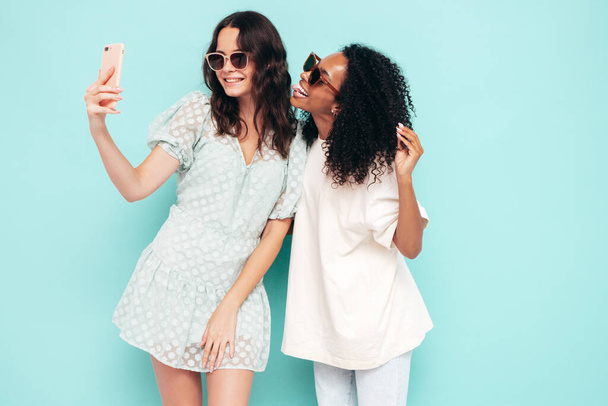 Two young beautiful smiling international hipster female in trendy summer clothes. Sexy carefree women posing near blue wall in studio. Positive models having fun in sunglasses. Taking selfie photos - Foto, Imagem