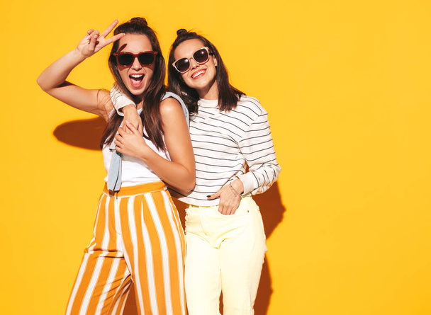 Two young beautiful smiling blond hipster female in trendy summer clothes. Sexy carefree women posing near yellow wall in studio. Positive models having fun. Cheerful and happy. In sunglasses - Foto, afbeelding