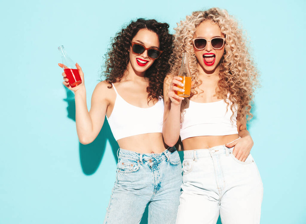 Two beautiful sexy smiling hipster women in summer clothes. Trendy models with afro curls hairstyle having fun in studio. Hot female isolated on blue. Drinking lemonade fro glass bottle. In sunglasses - Fotoğraf, Görsel