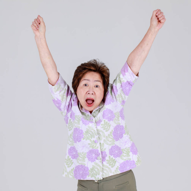 Mulher chinesa idosa engraçada agindo surpreendentemente levantando a mão para cima e apertando o punho com gritando em voz alta como emocionante para jogo maravilhoso e desfrutar de animar a concorrência como torcedor de torcedores sênior. - Foto, Imagem