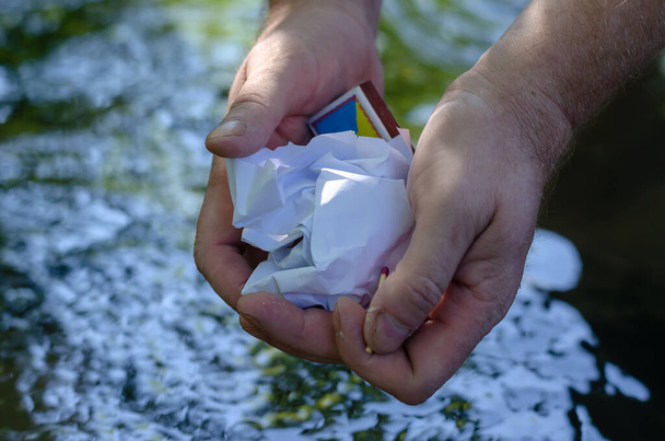 Twee mannen handen met een gerimpeld stuk papier en een doos lucifers. Een verkreukelde bal van wit papier in handen over het oppervlak van een rivier of beek. Selectieve focus. - Foto, afbeelding