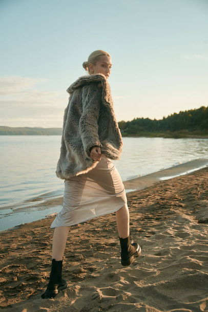 Fantastic blonde woman walking on the sand beach on a sea background. Beautiful tall model, slim figure. Silk grey dress, artificial fur coat. Black boots. Sunset. Blue sky. Romantic mood.   - Φωτογραφία, εικόνα