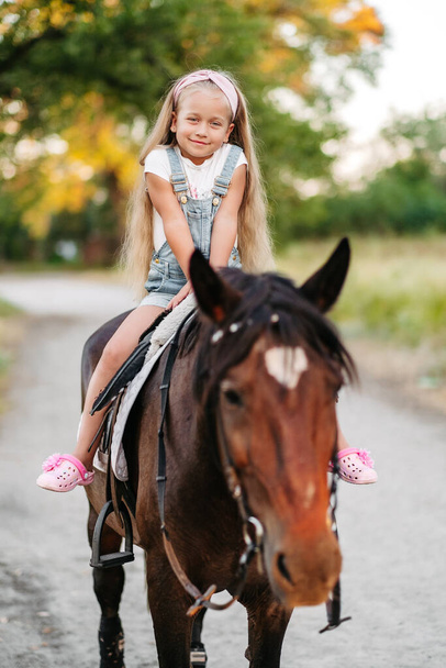 Little blonde girl with long hair rides a horse in the park at sunset in autumn. Autumn horse ride. Friendship of a girl and a horse. - Foto, Imagem