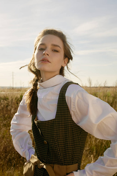 Portrait of young beautiful girl in village area. Big eyes and lips, long hair. Yellow field. Sun light, blue sky, autumn. Checkered dress, white shirt. - Фото, зображення