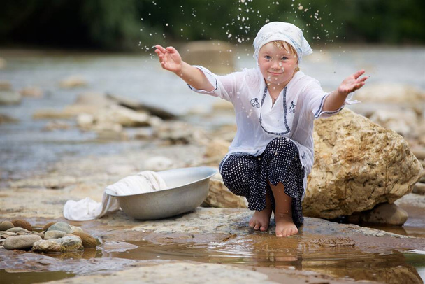 Dziewczyna w długiej wiosce vintage ubrania siedzi na brzegu rzeki i pluskania wodą. Rzeka z szybkim prądem w kraju z kamiennym brzegiem. - Zdjęcie, obraz