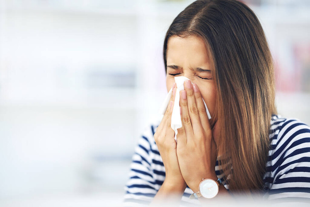 Its allergy season again. Shot of a young woman with allergies sneezing into a tissue at home. - Фото, изображение