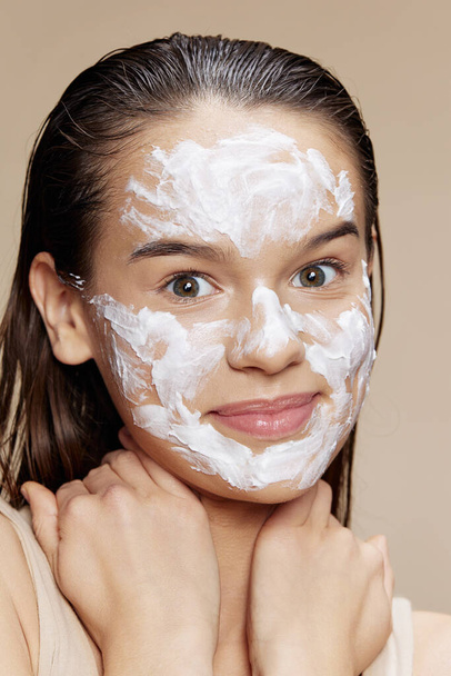 Mujer joven sonriente aplicando una mascarilla facial calmante cuidado de la piel fondo beige - Foto, imagen