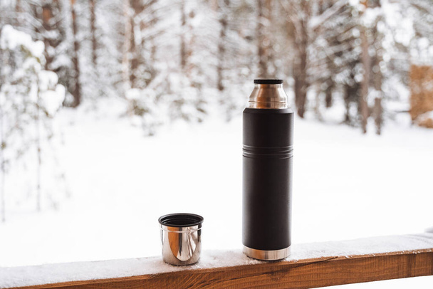 black matte thermos and mug with hot drink on window sill of the outdoor veranda. Hiking and picnic staff. Drinking warm tea or coffee in the winter frost. High quality photo - 写真・画像