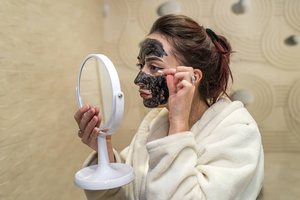 la fille dans la salle de bain se examine dans le miroir et corrige le masque de beauté nouvellement appliqué sur son visage. Concept de masque de beauté - Photo, image