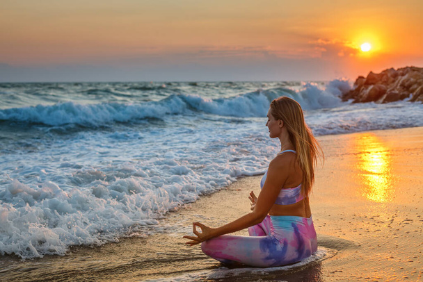 Woman with closed eyes sitting in a lotus position on the beach during sunset. Inner harmony and self body care. - Fotoğraf, Görsel