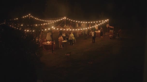 Widok z powietrza na nowożeńców tańczących wśród gości na trawniku z układanymi stołami i rzędami girland z płonącymi żarówkami - Materiał filmowy, wideo