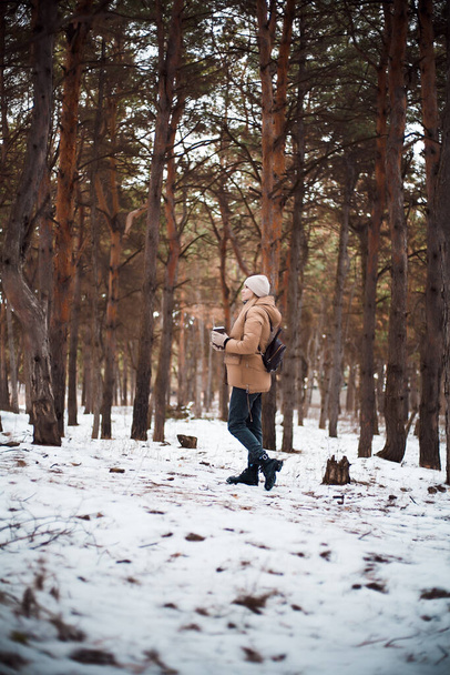 Ένα ελκυστικό κορίτσι με ξανθά μαλλιά και ένα φλιτζάνι ζεστό ρόφημα περπατά στο δάσος. - Φωτογραφία, εικόνα