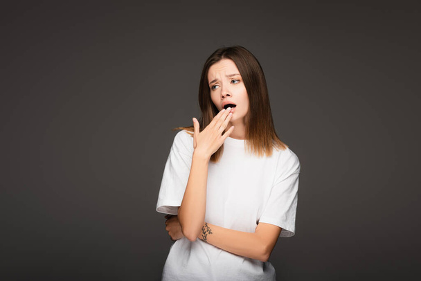 bored woman in white t-shirt covering mouth with hand while yawning isolated on dark grey - Photo, Image