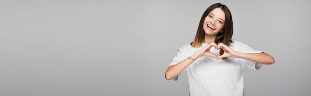 happy brunette woman showing heart sign with hands isolated on grey, banner - Fotografie, Obrázek