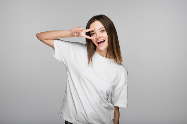 excited brunette woman showing victory sign near face isolated on grey - Fotó, kép