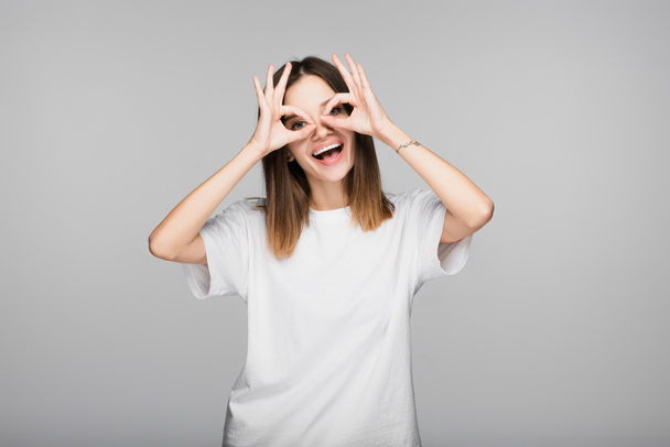 vrolijke vrouw in wit t-shirt met goede tekenen in de buurt van ogen geïsoleerd op grijs - Foto, afbeelding