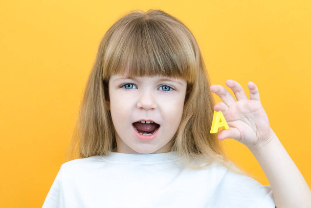 speech therapy. Toddler girl holding the letter A in her hands. Classes with a speech therapist. Girl on isolated yellow background - 写真・画像