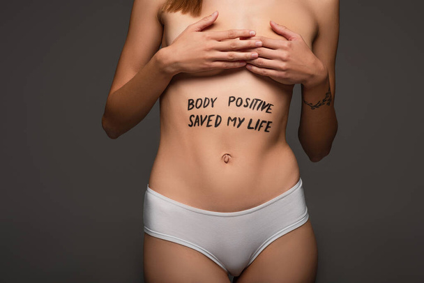 partial view of woman with body positive saved my life lettering on belly covering breast with hands isolated on dark grey - Photo, Image