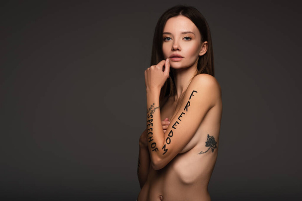 mujer desnuda y tatuada con frase de libertad de expresión escrita en un brazo aislado en gris oscuro - Foto, Imagen