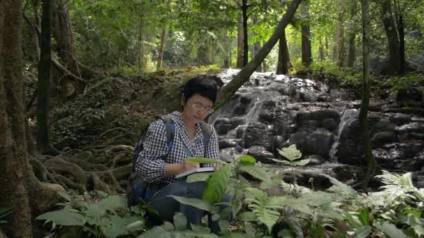 Asyalı kadın botanik araştırmacısı şelale etrafında yetişen bitkilerin özelliklerini inceliyor ve öğreniyor. Orman doğası ve fotosentezin biyolojik süreci. Deftere yazma ve çizme. - Video, Çekim