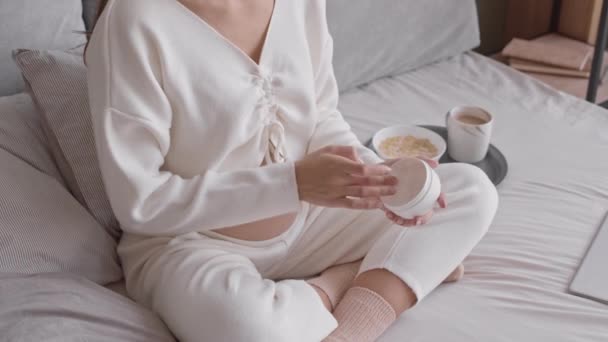 Tiro lento recortado de una joven embarazada irreconocible cuidando su piel durante el embarazo, aplicando crema de estrías en su vientre, sentada en la cama en casa - Metraje, vídeo