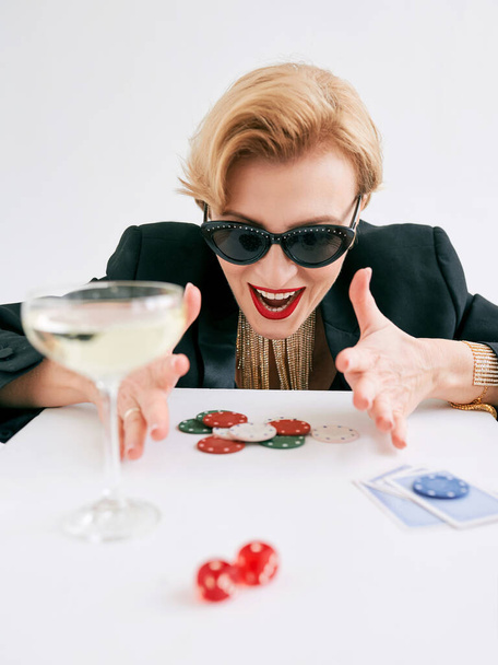 reife, stylische Frau in schwarzem Smoking und Sonnenbrille, die glücklich ist, im Casino zu gewinnen. Glücksspiel, Mode, Hobbykonzept. - Foto, Bild