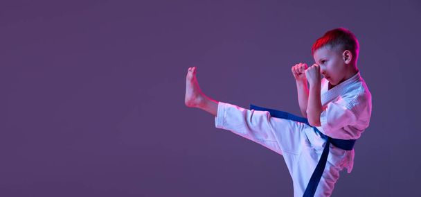 Portrait of sportive kid, male taekwondo, karate athletes in doboks doing basic movements isolated on purple background in neon. Concept of sport, martial arts - Foto, Bild