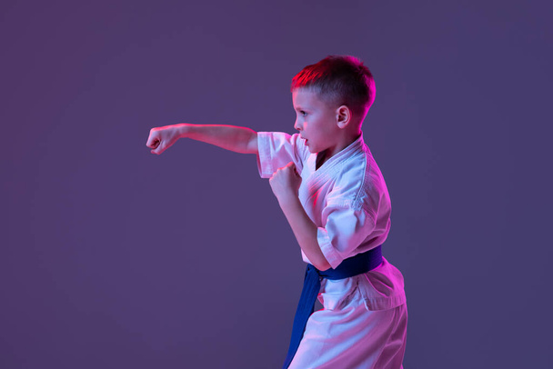 Ritratto dinamico di bambino, giovane maschio taekwondo, atleti di karate in dobok che fanno movimenti di base isolati su sfondo viola al neon. Concetto di sport, arti marziali - Foto, immagini