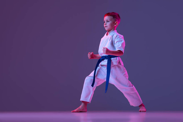 Dynamic portrait of kid, young male taekwondo, karate athletes in doboks doing basic movements isolated on purple background in neon. Concept of sport, martial arts - Valokuva, kuva