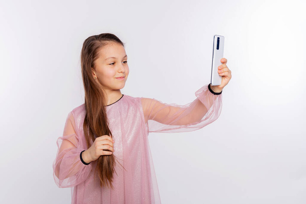 Παιδί κορίτσι φορώντας ροζ φόρεμα φυσάει αέρα φιλί στην κάμερα του smartphone και παίρνει selfie σε λευκό φόντο. Ελεύθερος χώρος για το κείμενο σας - Φωτογραφία, εικόνα