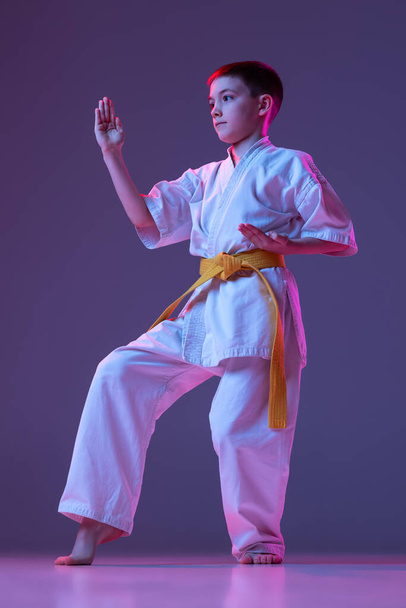 Studio shot of sportive kid, male taekwondo, karate athletes in doboks posing isolated on purple background in neon. Concept of sport, martial arts - Foto, Imagen