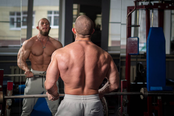 Man admires his reflection in mirror of the fitness room. Bodybuilder holds in his hands and trains with barbell in the gym. Pumped up guy show muscles. Concept of an active healthy lifestyle - Photo, Image
