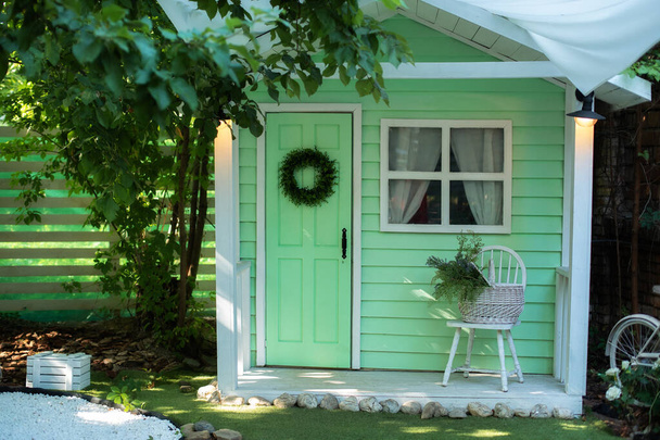 Fasáda zelený dřevěný dům zdobený pro jarní prázdniny. Exteriér Dřevěná veranda domů se zahradním nábytkem. Interiér útulný veranda dům s židlí a košíkem květin. Dekor domácí venkovní letní zahrada - Fotografie, Obrázek