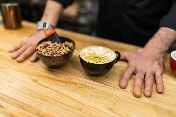 unrecognizable barista, showing a pistachio latte on the wooden bar of the coffee shop next to a bowl of pistachios. - Foto, Imagen